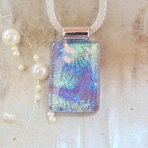 May include: A rectangular dichroic glass pendant with a silver chain. The pendant displays a blend of blue, purple, and green hues with a textured, iridescent pattern. The necklace is paired with a strand of white pearls.