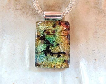 May include: A rectangular pendant with a dichroic glass design in shades of green, gold, and black, suspended from a silver chain. The glass has a textured appearance with dark, organic-looking inclusions. The pendant is approximately 2.5 cm long.