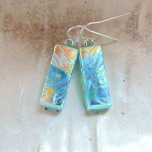 Puede incluir: Un par de pendientes rectangulares con un diseño de vidrio dicróico. Los pendientes presentan una combinación de colores azul, dorado y verde. Los pendientes tienen ganchos para las orejas de color plateado.