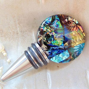 May include: A dichroic glass bottle stopper with a stainless steel base. The round top features a mosaic of colors including blue, green, gold, and red. The stopper has a silver base with black rubber rings.