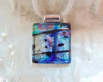 May include: A square dichroic glass pendant necklace with a silver chain. The pendant features a vibrant mix of colors, including blue, green, purple, and silver, with black accents. The necklace is a piece of jewelry.
