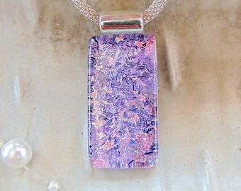 May include: A rectangular pendant with a shimmering, textured surface in shades of purple, pink, and blue. The pendant is suspended from a silver chain. A small pearl necklace is visible in the lower left corner.