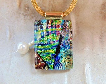 May include: A rectangular dichroic glass pendant featuring a vibrant, multi-coloured pattern of green, blue, and gold metallic flecks. The pendant is suspended from a gold-toned mesh chain and is positioned next to a single white pearl bead.