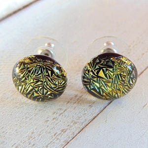 May include: A pair of round stud earrings. Each earring features a dark base with a gold and green geometric pattern. The earrings have a post and backing for secure wear. The earrings are displayed on a white wooden surface.