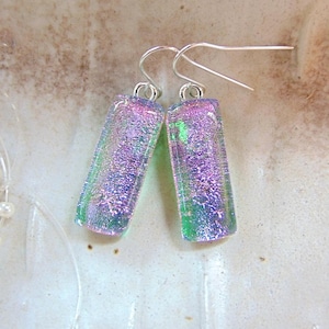 May include: A pair of rectangular dichroic glass earrings featuring a shimmering blend of iridescent purple, pink, and green hues. Each glass piece is suspended from a simple silver-toned metal hook, creating a reflective and textured surface.