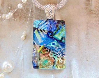 May include: A rectangular dichroic glass pendant featuring a vibrant abstract pattern of blue, green, and gold tones. The pendant is suspended from a silver-toned woven mesh chain, held by a textured, chevron-patterned silver-toned metal bail.