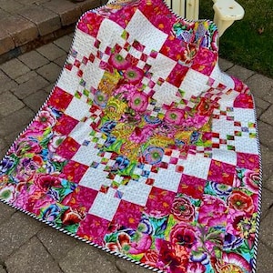 May include: A pink and white quilt with a floral pattern. The quilt is folded and sitting on a stone patio.