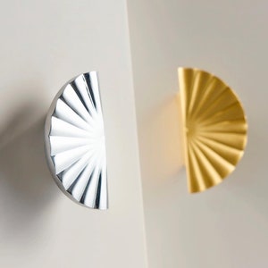 Op de afbeelding: Twee decoratieve kastdeurgrepen, een zilveren en een gouden, in de vorm van een halve cirkel met een waaiervormig ontwerp.