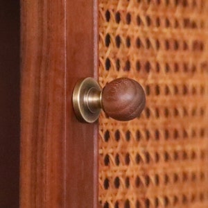 Pode incluir: Close-up de uma porta de armário de madeira com uma maçaneta de latão e um painel de vime tecido. A maçaneta tem um detalhe em madeira escura.