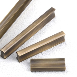 以下が含まれることがあります： アンティーク真鍮のキャビネットプルハンドルのクローズアップ。プルハンドルは長方形で、縁が湾曲し、中央がくぼんでいます。仕上げは暖かく、古びた真鍮色です。プルハンドルは、質感のある白い表面に配置されています。