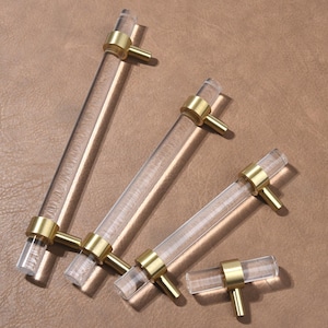 May include: Clear acrylic cabinet handles with brass-coloured fittings. The handles are cylindrical and feature a contemporary design. The brass fittings are circular and connect the handle to the cabinet. The handles are of different lengths.