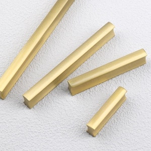 May include: A collection of gold-toned cabinet pulls in various sizes. The pulls have a rectangular shape with a slightly curved handle, and are arranged on a light-colored surface. The hardware is likely for furniture or cabinetry.