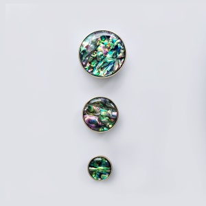Op de afbeelding: Drie ronde, iriserende parelmoer knoppen met een gouden rand. De knoppen zijn verticaal gerangschikt in aflopende volgorde.