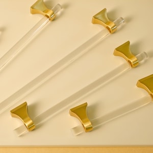 May include: Six gold and clear acrylic cabinet pulls with a simple, modern design. The pulls are arranged in a fan shape on a light beige surface.
