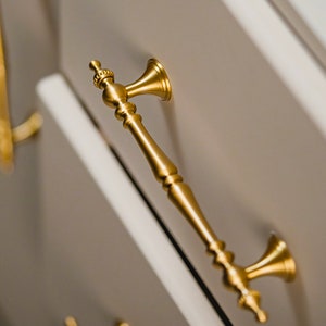 May include: Gold cabinet hardware with a decorative design. The handle is attached to a white cabinet door.