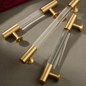 May include: Three cabinet pulls with a clear acrylic center and gold-toned metal ends and handles. The pulls are arranged diagonally across a gray surface, showcasing their design and hardware details. The hardware is designed for furniture.