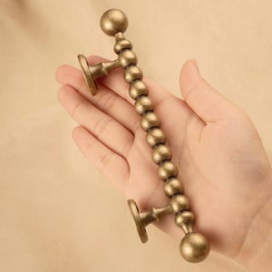 May include: A bronze-colored cabinet pull handle with a beaded design. The handle has a curved shape with a ball at each end and circular mounting plates. The handle is held in a hand.