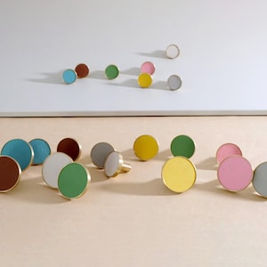 Op de afbeelding: Assortiment van ronde, kleurrijke, met leer beklede knoppen met een messing rand. De knoppen zijn in twee rijen op een lichtbruin oppervlak gerangschikt.