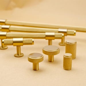 Puede incluir: Un juego de herrajes para armarios de color dorado, que incluye pomos y tiradores. Los pomos son redondos con una superficie texturizada, y los tiradores tienen forma de T con una barra texturizada. El herraje está hecho de metal y tiene un acabado pulido.