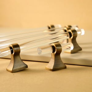 May include: Four clear glass cabinet pulls with antique brass bases. The pulls are cylindrical and have a smooth, polished finish.