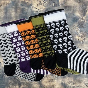 Könnte beinhalten: Fünf gestrickte Strümpfe mit verschiedenen Halloween-Motiven. Die Strümpfe sind schwarz, weiß, orange, grün und lila mit verschiedenen Designs, darunter Fledermäuse, Geister, Kürbisse, Katzen und Schädel.