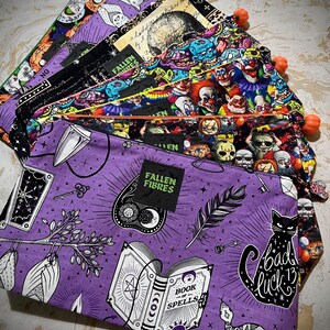 May include: A stack of purple, black, and green zippered pouches with various Halloween-themed prints. The top pouch features a black cat with the text "bad luck 13" and a book with the text "Book of Spells". The pouch also has a green tag with the text "Fallen Fibres".