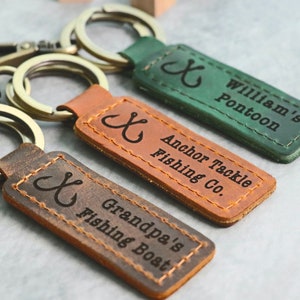 May include: Three leather keychains with a fishing hook design and personalised text. The keychains are brown, green and dark brown. The text on the keychains reads "Anchor Tackle Fishing Co.", "William's Pontoon", and "Grandpa's Fishing Boat".