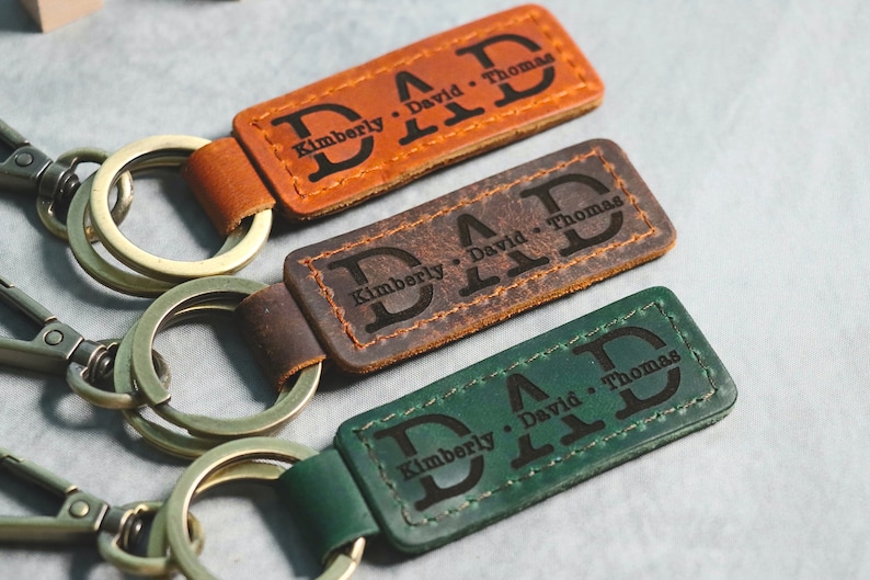 May include: Three leather keychains with metal rings. Each keychain has the word "DAD" engraved in large letters, with the first letter of the word "DAD" being a monogrammed initial. The keychains are brown, dark brown, and green.