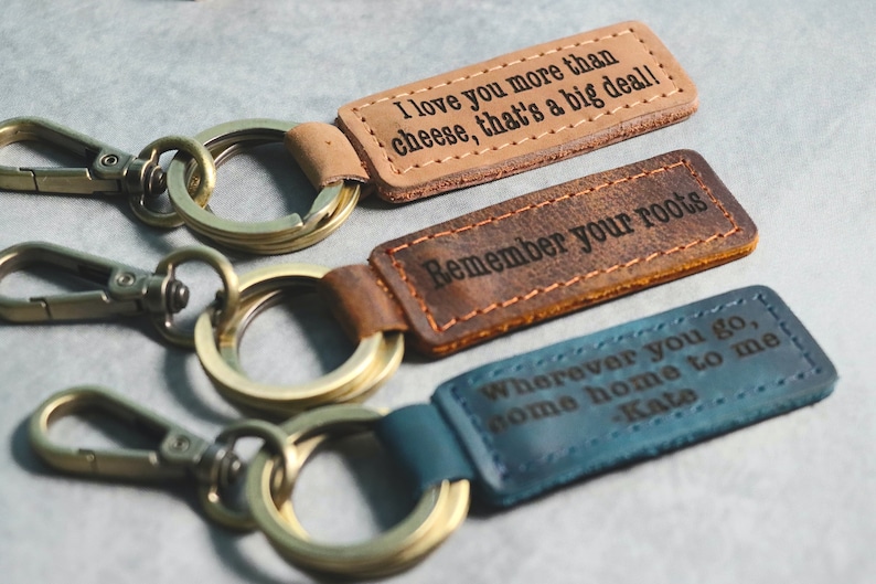 May include: Three leather keychains with different engraved messages. The first keychain is brown and says "I love you more than cheese, that's a big deal!" The second keychain is brown and says "Remember your roots". The third keychain is blue and says "Wherever you go, home home to me - Kate".