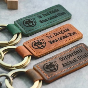 May include: Three leather keychains with metal rings and a lobster clasp. Each keychain has a different colour and features a paw print with a stethoscope and the name of a veterinary clinic. The keychains read "Dr. Paul Fidelis Mena Animal Clinic", "Dr. Douglass Mena Animal Clinic", and "Copperfield Animal Clinic".