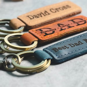 May include: Three leather keychains with metal rings. The top keychain is tan with the text "David Cross" engraved on it. The middle keychain is brown with the text "Kimberly David Thomas" engraved on it. The bottom keychain is blue with the text "Best Dad Ever!" engraved on it.