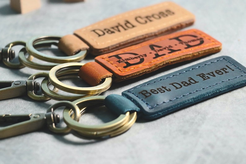 May include: Three leather keychains with metal rings. The top keychain is tan with the text "David Cross" engraved on it. The middle keychain is brown with the text "Kimberly David Thomas" engraved on it. The bottom keychain is blue with the text "Best Dad Ever!" engraved on it.