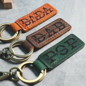 May include: Three leather keychains with metal rings and clasps. Each keychain has a different color: brown, dark brown, and green. The keychains are personalized with the text "DAD" and the names "Kimberly, David, Thomas".