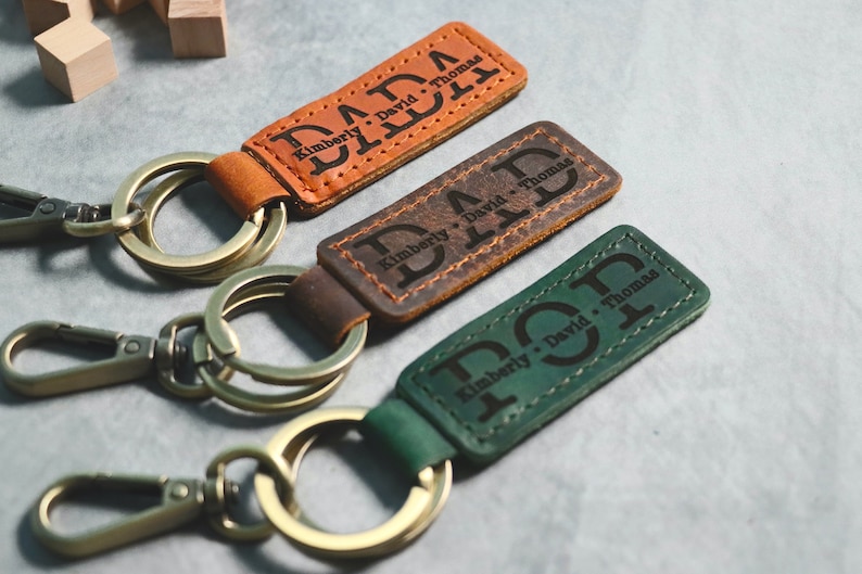 May include: Three leather keychains with metal rings and clasps. Each keychain has a different color: brown, dark brown, and green. The keychains are personalized with the text "DAD" and the names "Kimberly, David, Thomas".