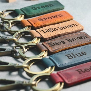 May include: Six leather keychains with metal rings and clasps. The keychains are in different colors: green, brown, light brown, dark brown, blue, and red. Each keychain has a leather tag with the color name embossed on it.