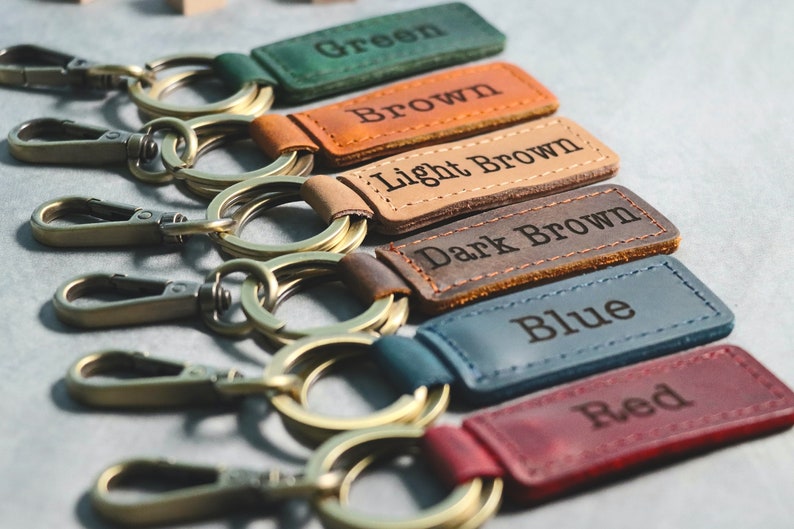 May include: Six leather keychains with metal rings and clasps. The keychains are in different colors: green, brown, light brown, dark brown, blue, and red. Each keychain has a leather tag with the color name embossed on it.