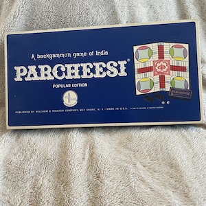 Puede incluir: Una caja azul del juego de mesa Parcheesi. La caja presenta el nombre del juego en letras blancas grandes, con el texto "A backgammon game of India" encima. La caja también incluye el texto "Popular Edition" y una ilustración del tablero de juego.