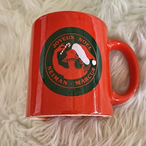Puede incluir: Una taza de cerámica roja con asa. La taza presenta un emblema circular con las palabras "Joyeux Noel" y "Neiman Marcus" en verde. El emblema incluye una figura de Papá Noel y un bastón de caramelo.