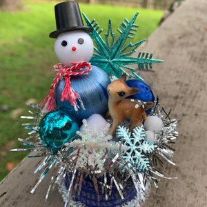 Puede incluir: Una decoración navideña azul y blanca con un muñeco de nieve con un sombrero de copa negro, una figurita de ciervo azul y blanca y un adorno de copo de nieve azul y blanco. La decoración está rodeada de oropel plateado y copos de nieve blancos.