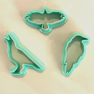 May include: Three teal-colored bird-shaped cookie cutters. The cutters are arranged on a beige square, set against a backdrop of a blue and white patterned surface. The bird shapes include a flying bird, a perched bird, and a bird profile.
