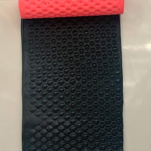 May include: A textured black material is partially rolled out on a cream-colored rectangular surface. A bright pink roller with a honeycomb pattern sits atop the black material. The background features a teal and white floral pattern.