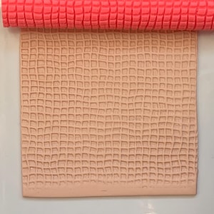 Puede incluir: Un rodillo texturizado de color coral descansa sobre un cuadrado beige con un patrón de cuadrícula, ambos sobre una baldosa cuadrada blanca. Los elementos están colocados sobre un fondo estampado con diseños florales turquesa y blancos. El rodillo parece ser para manualidades.