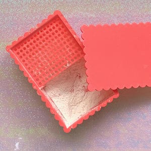 Peut inclure: Un boîtier compact corail ouvert avec un bord festonné, révélant une poudre fine et claire. Le couvercle est légèrement entrouvert et l'intérieur présente un motif perforé. Le fond est une surface chatoyante et irisée.