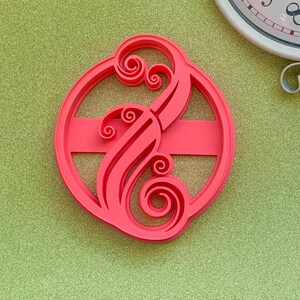 May include: A bright pink, circular cookie cutter with a decorative, swirling design. The cutter features a stylized letter "B" with ornate curls and a horizontal bar. The background is a textured, light green surface, with a white clock in the background.