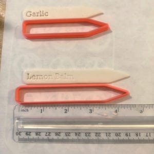 Może przedstawiać: Dwa białe znaczniki do roślin z wytłoczonym napisem Garlic i Lemon Balm, w parze z dwiema pomarańczowymi plastikowymi foremkami. Przezroczysta linijka na dole pokazuje, że znaczniki mają około 11,5 cm długości całkowitej.