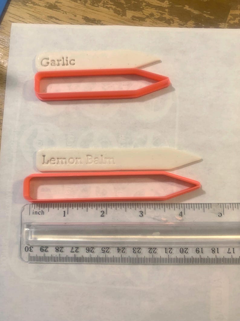 Może przedstawiać: Dwa białe znaczniki do roślin z wytłoczonym napisem Garlic i Lemon Balm, w parze z dwiema pomarańczowymi plastikowymi foremkami. Przezroczysta linijka na dole pokazuje, że znaczniki mają około 11,5 cm długości całkowitej.