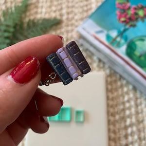 May include: A miniature charm featuring three stacked books in shades of purple and navy blue with gold accents. The charm is attached to a silver chain. The background includes a book and small green objects.