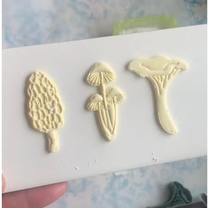 May include: Three off-white mushroom designs are displayed on a white surface. The mushrooms include a morel, a cluster of two, and a chanterelle. The designs are likely for crafting or decorative purposes, showcasing a nature-inspired theme.