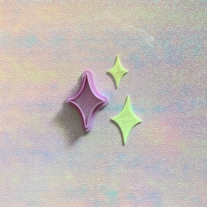Puede incluir: Tres objetos en forma de estrella. Uno es un cortador de galletas de color lila claro, y los otros dos son estrellas de color verde lima. El fondo es una superficie texturizada e iridiscente con colores pastel.