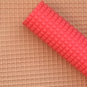 May include: A bright red, textured, cylindrical object rests diagonally on a tan, grid-patterned surface. The red object has a repeating, scale-like pattern. The background surface has a repeating square pattern.
